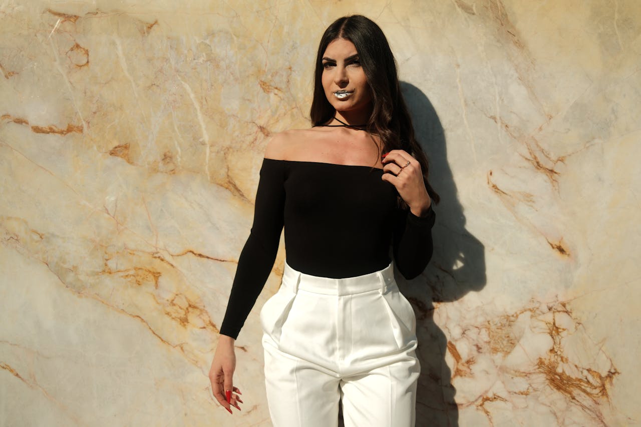 services-02 Stylish woman posing in a black off-shoulder top and white pants against a marble wall.