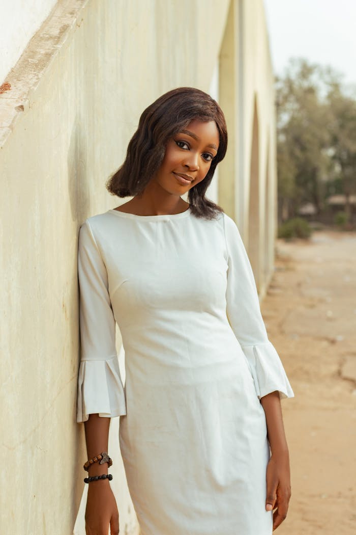 Portrait of a woman in white dress leaning against a wall with graceful elegance in a sunlit outdoor setting.